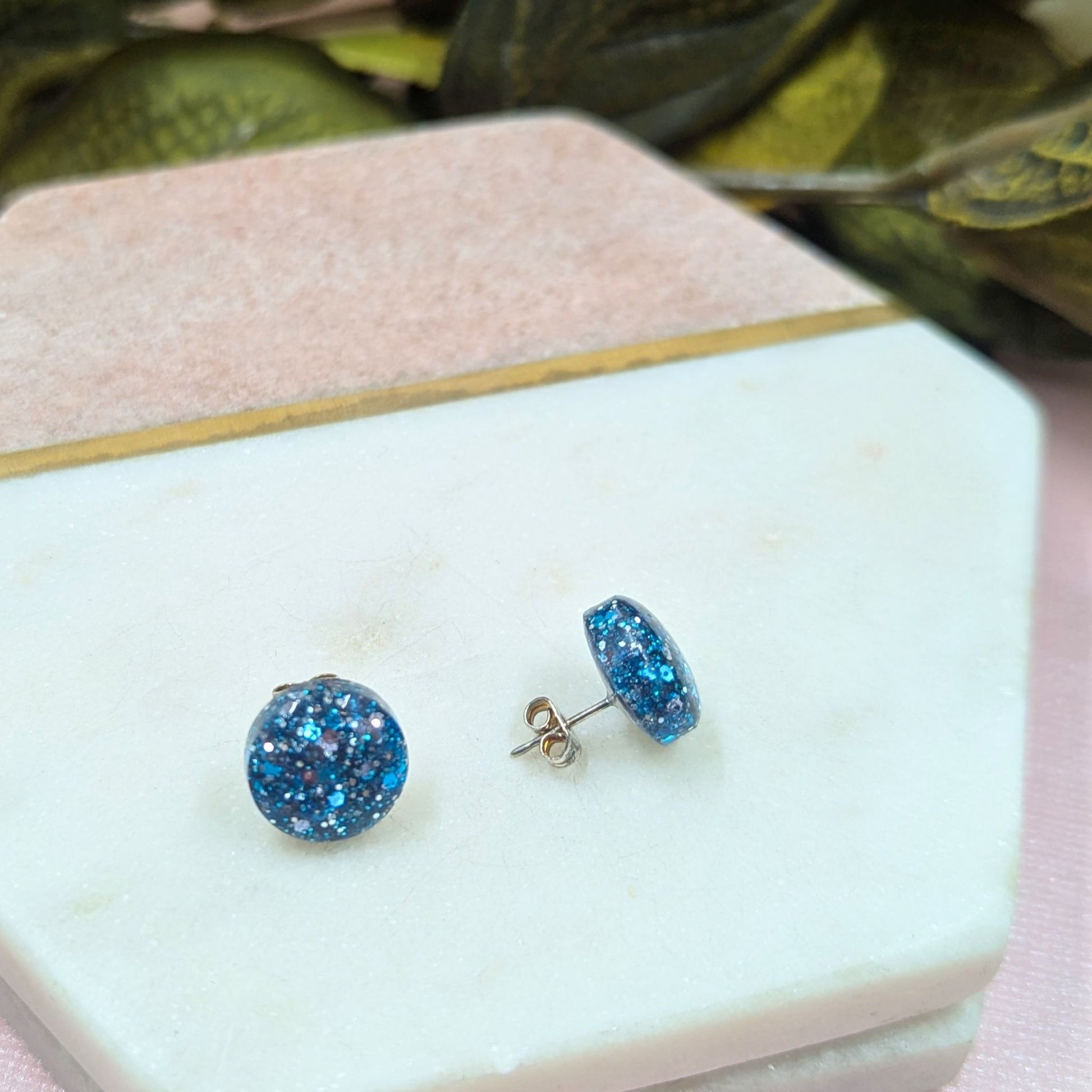 Blue glittery stud earrings on a white marble surface with green leaves in the background