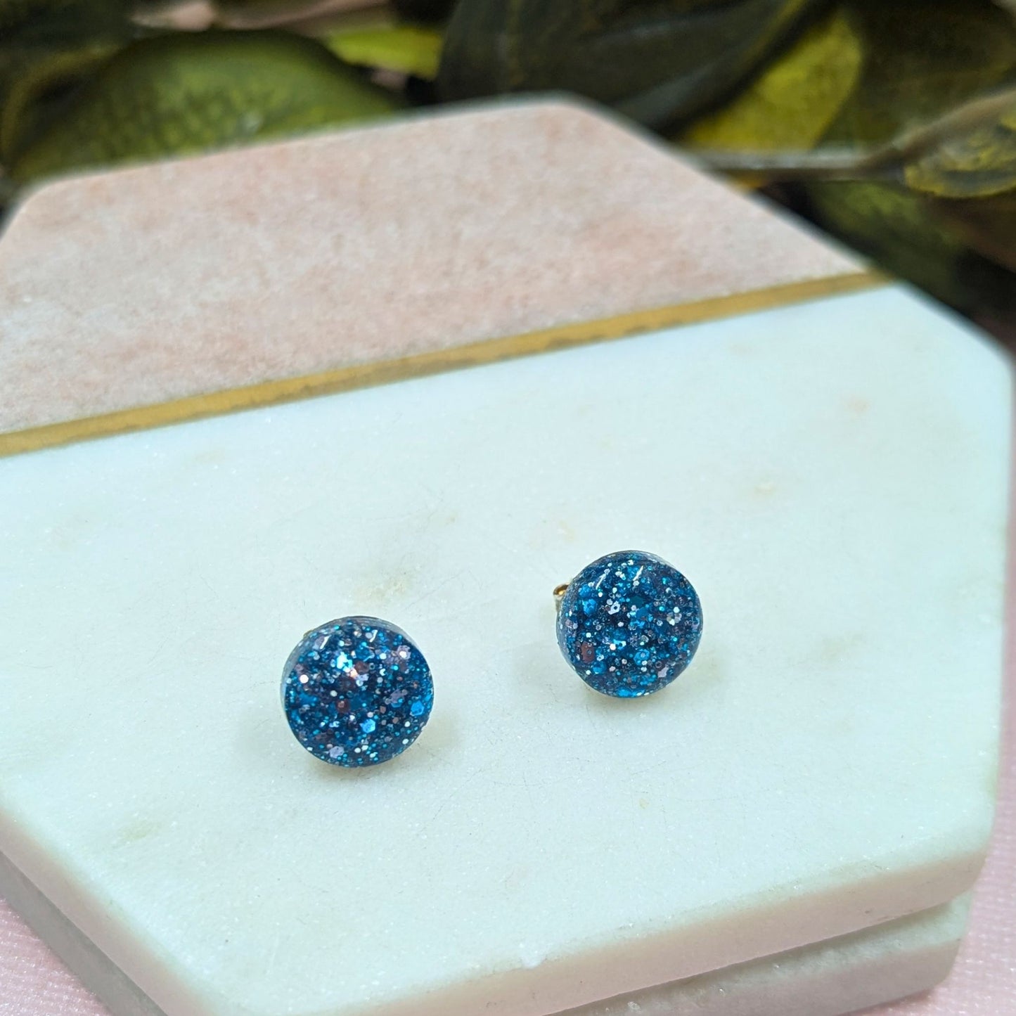 Blue glittery stud earrings on a white jewelry box with green leaves in the background