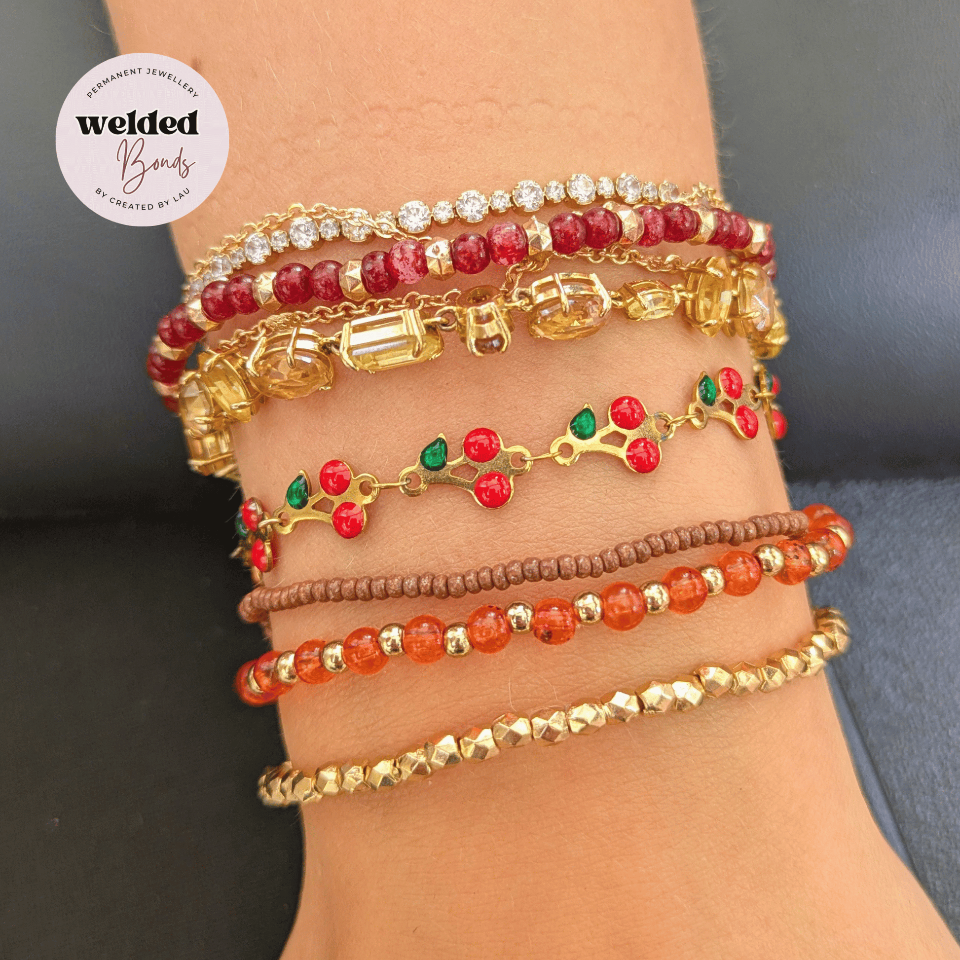 A stack of five multi-material gold-plated woven and beaded bracelets displayed on a person's wrist along side  our Bonn Cherry Chain Welded Bracelet. The bracelets feature red, brown, and orange beads, clear rhinestones, and small green and red accents.