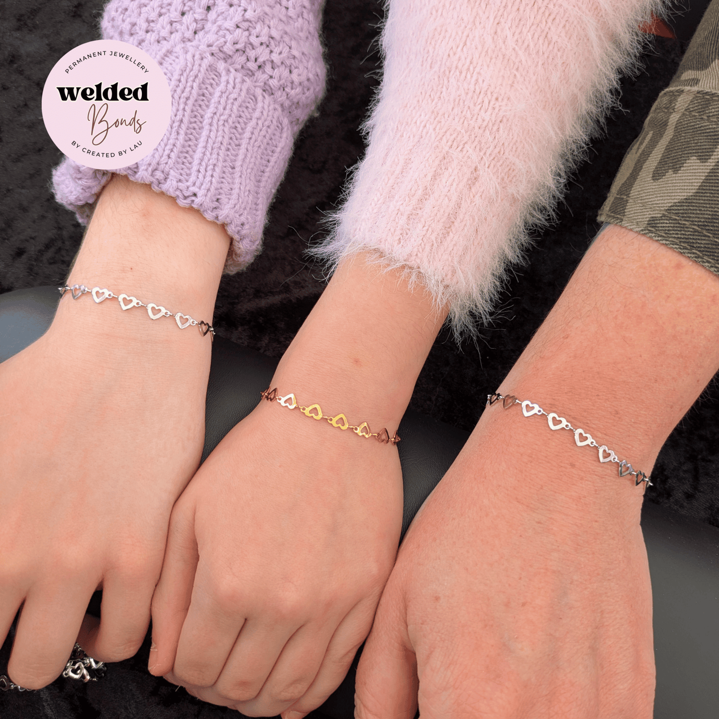 Three delicate heart-link bracelets shown on three wrists, with a white circular logo in the top left reading 'welded Bands'. The bracelets feature Welded Bonds "Salzburg" Stainless Steel Flat Heart Chain in silver and gold metal tones, worn over purple, pink, and camouflage knit sleeves.