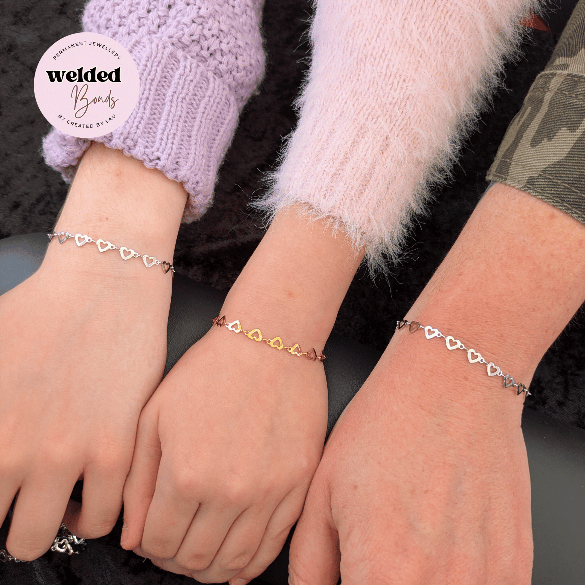 Three delicate heart-link bracelets shown on three wrists, with a white circular logo in the top left reading 'welded Bands'. The bracelets feature Welded Bonds "Salzburg" Stainless Steel Flat Heart Chain in silver and gold metal tones, worn over purple, pink, and camouflage knit sleeves.