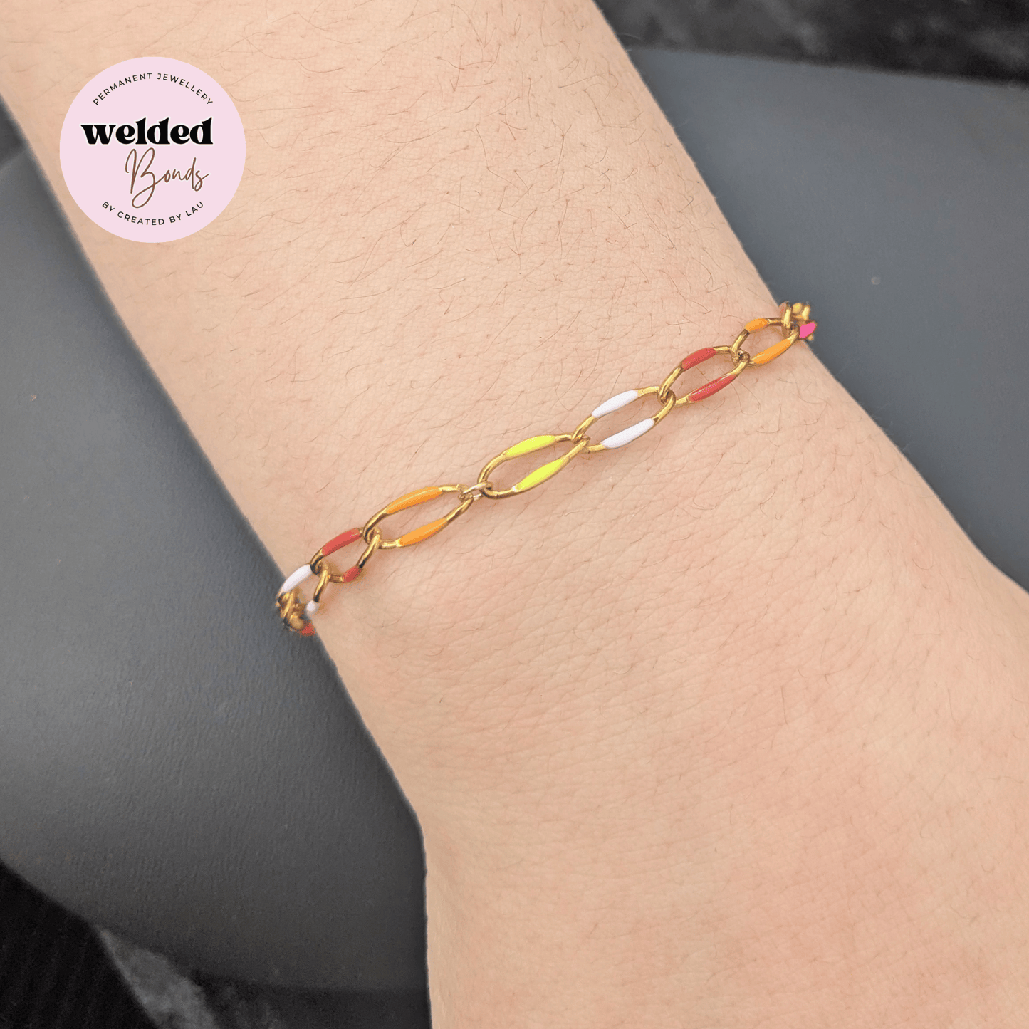 Our "Kingston" Stainless Steel Enamel Chain bracelet with alternating translucent orange, yellow, and white oval links, shown worn on a person's wrist. A circular logo for 'welded Bands' is visible in the top-left corner.