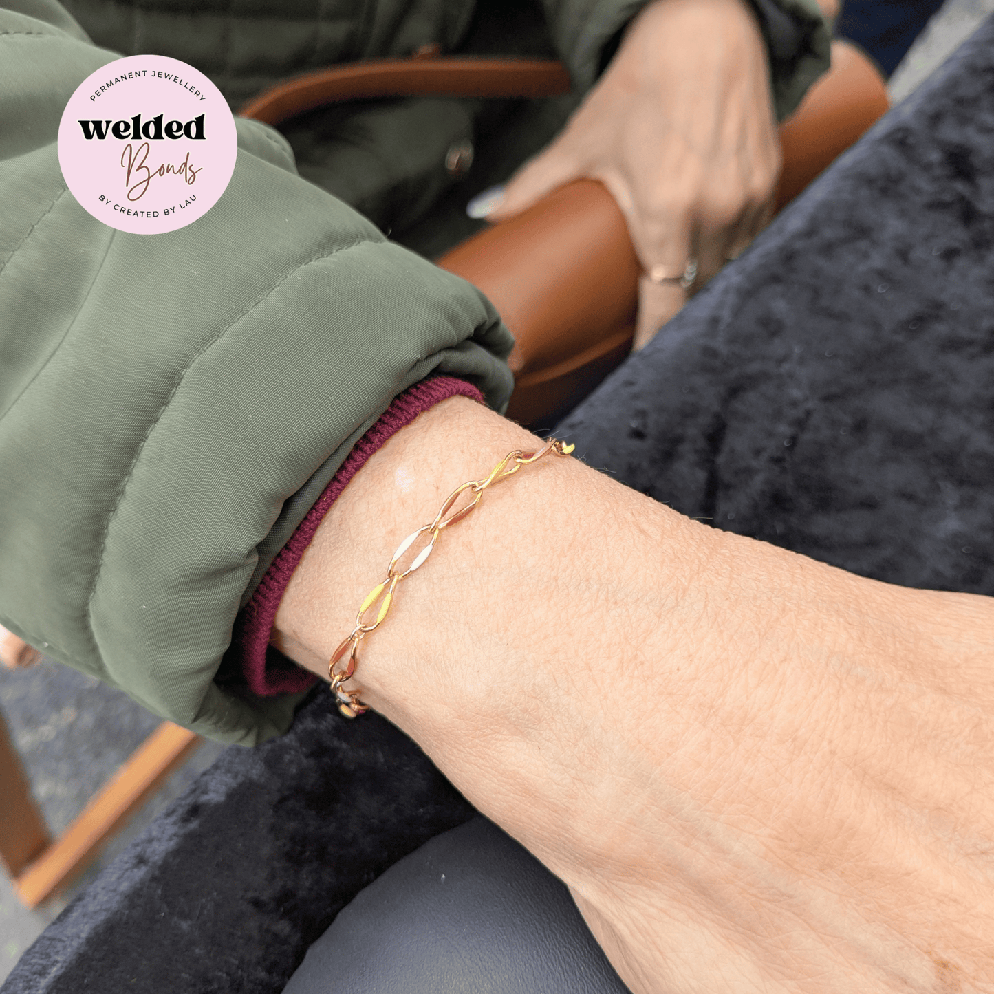 A close-up of a person's wrist wearing our Welded Bonds "Kingston" Stainless Steel Enamel Chain, styled with an olive green sleeve and a burgundy ribbed cuff. A circular logo for 'Welded Bands' is visible in the top left corner.