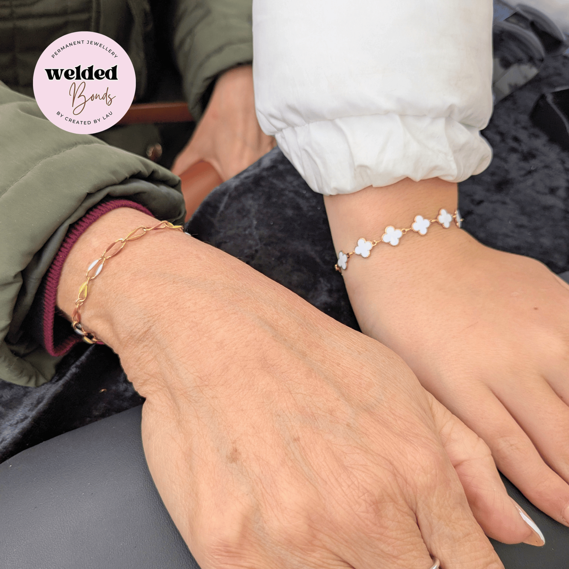 Two delicate gold bracelets shown on a person's wrist and hand. One bracelet is our Welded Bonds "Kingston" Stainless Steel Enamel Chain with multi coloured enamal, and the other is our Welded Bonds "Lyon" Stainless Steel Enamel Clover Chain in white.