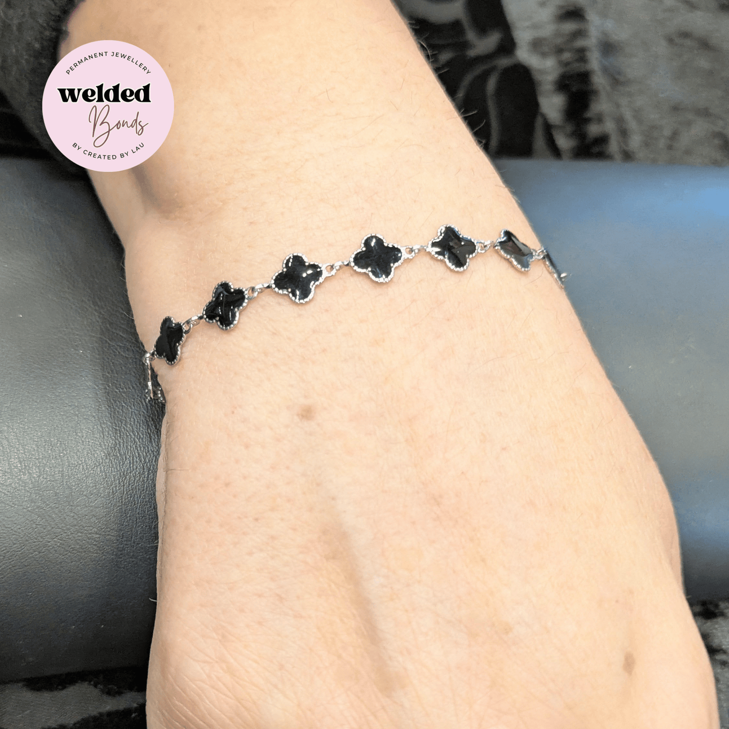 A close-up of a person's wrist wearing a silver-toned bracelet with five black clover motifs, with a circular 'Welded Bonds by Created By Lau' logo overlay in the top left corner.