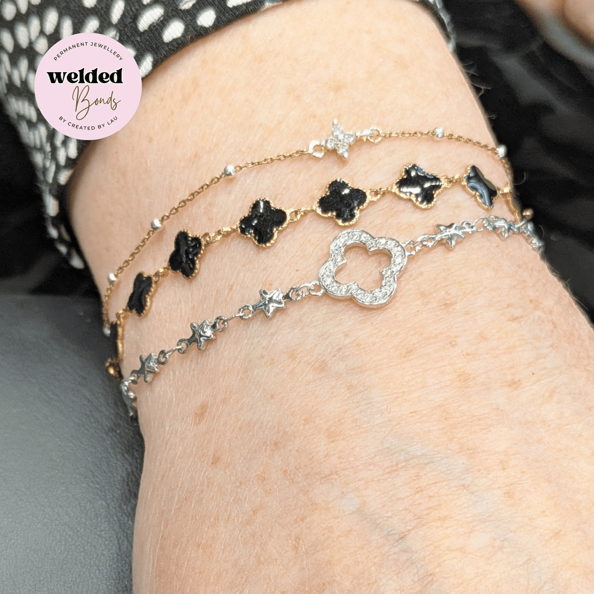 A close-up of a person's wrist wearing three delicate layered bracelets: a gold-tone bracelet with black clover motifs, a silver-tone bracelet with black clover motifs, and a silver-tone bracelet with a central diamond-encrusted clover motif and star links. A pink circular badge in the top left reads 'Welded Bonds by Created by Lau'.