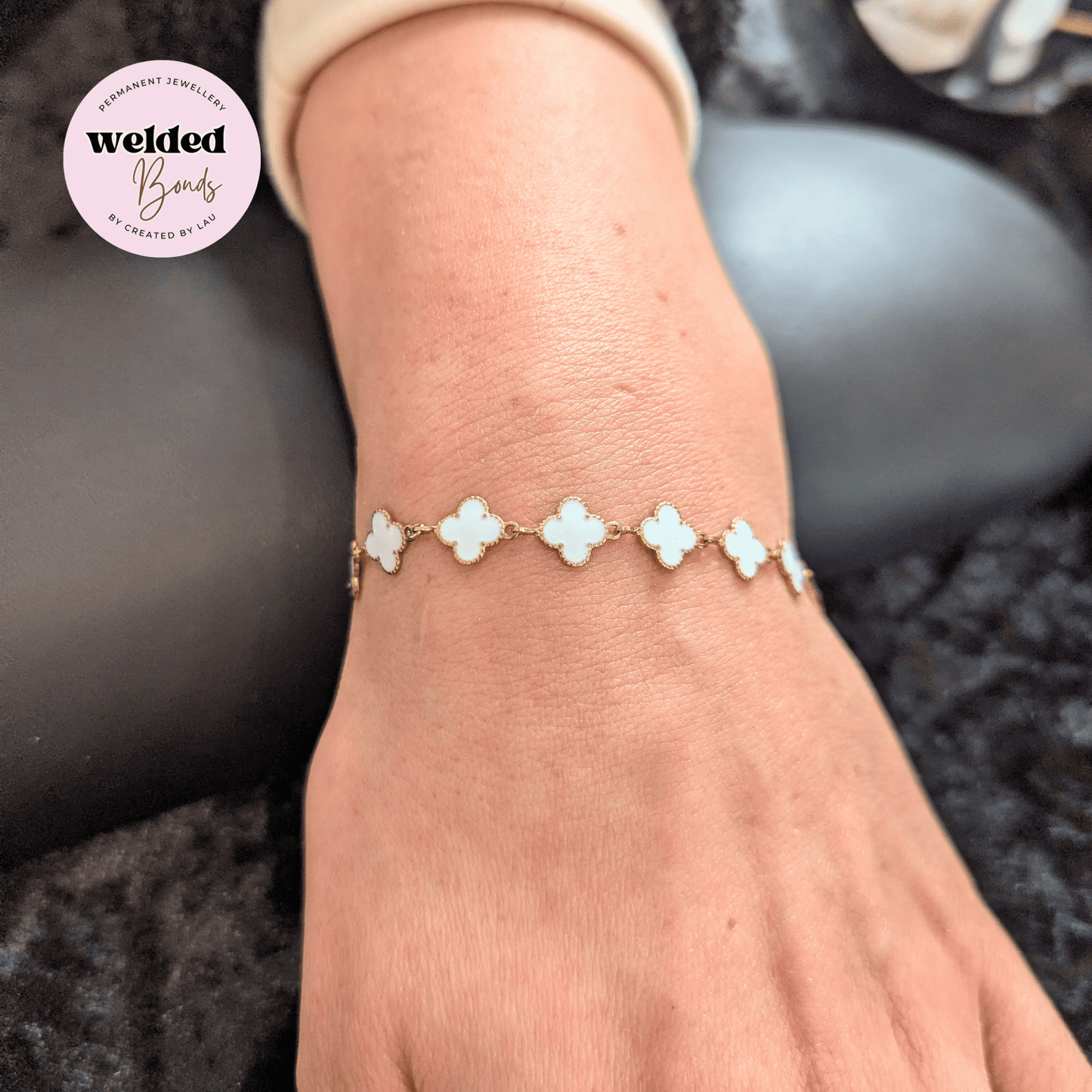 A close-up of a person's wrist wearing a delicate bracelet with five white clover-shaped links connected by gold-toned metal, shown against a dark textured background. A circular 'welded Bonds' brand logo is visible in the top-left corner.