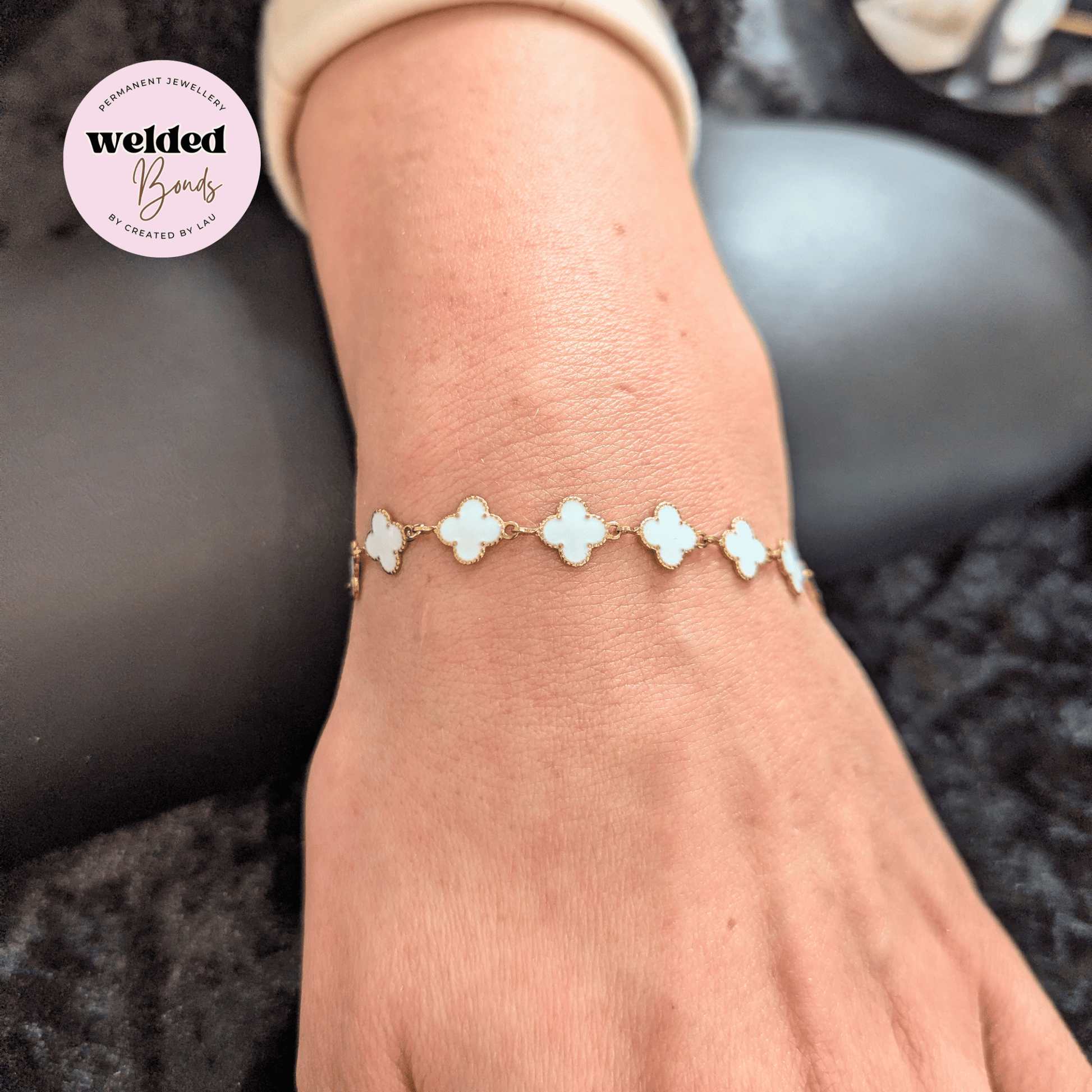 A close-up of a person's wrist wearing a delicate bracelet with five white clover-shaped links connected by gold-toned metal, shown against a dark textured background. A circular 'welded Bonds' brand logo is visible in the top-left corner.