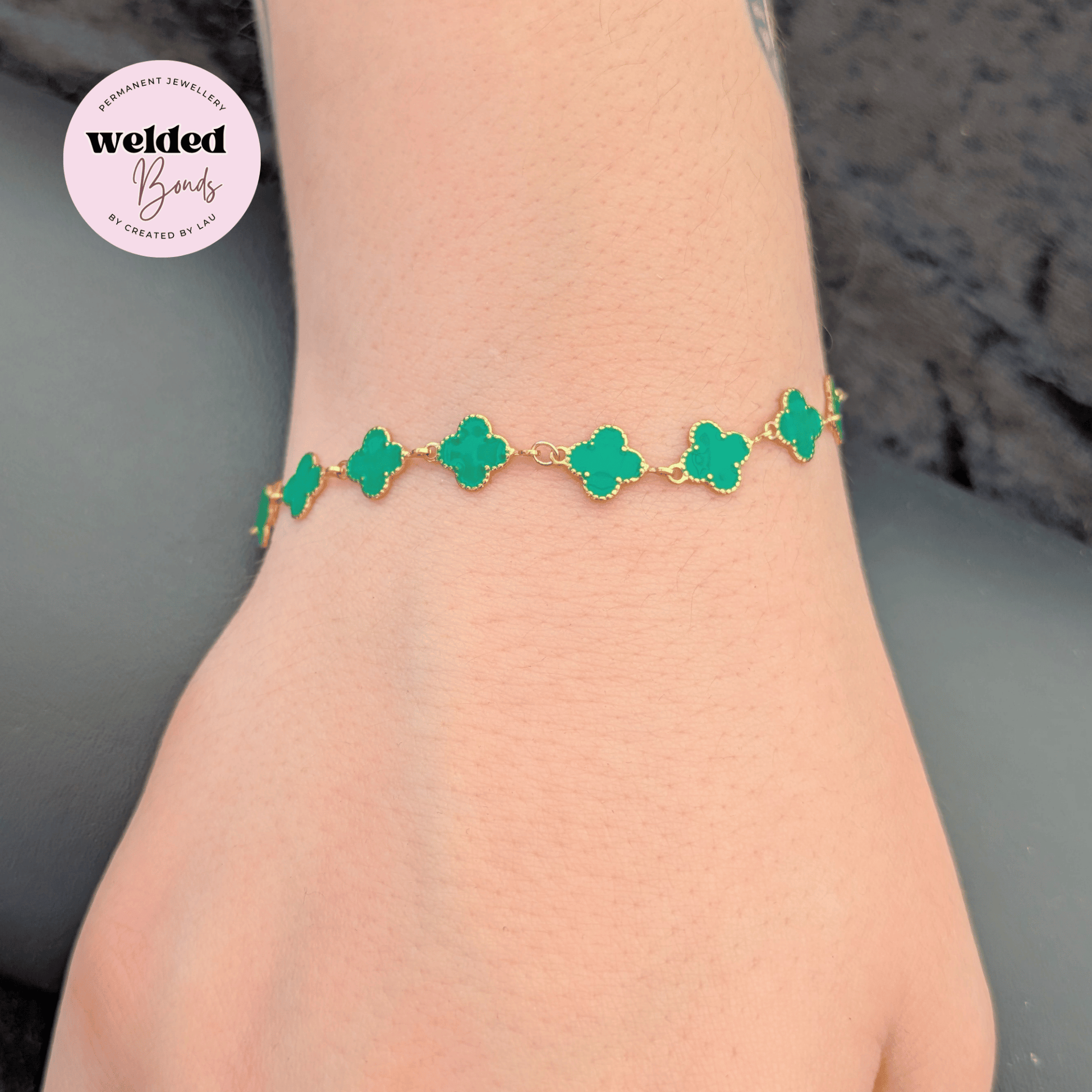 A gold chain bracelet with green clover-shaped links worn on a person's wrist, shown against a neutral grey background with a circular 'Welded Bonds' badge in the top left corner.