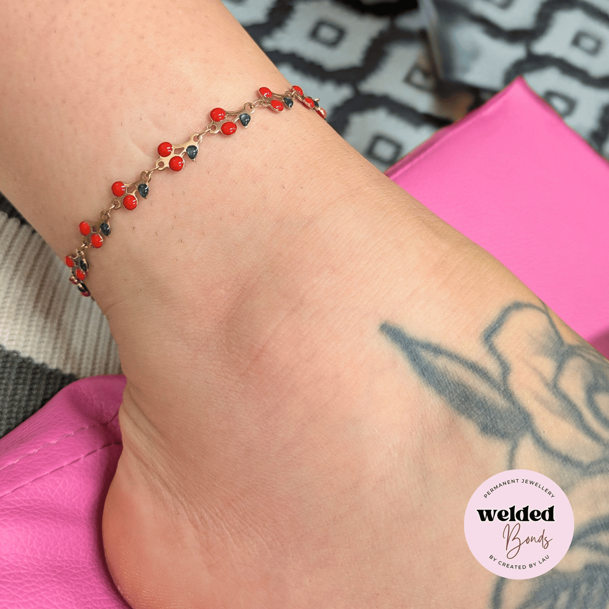 A close-up of a person's ankle wearing a delicate gold chain bracelet adorned with small red and green enamal beads. The person's foot has a grey tattoo, and a pink surface is visible in the background.