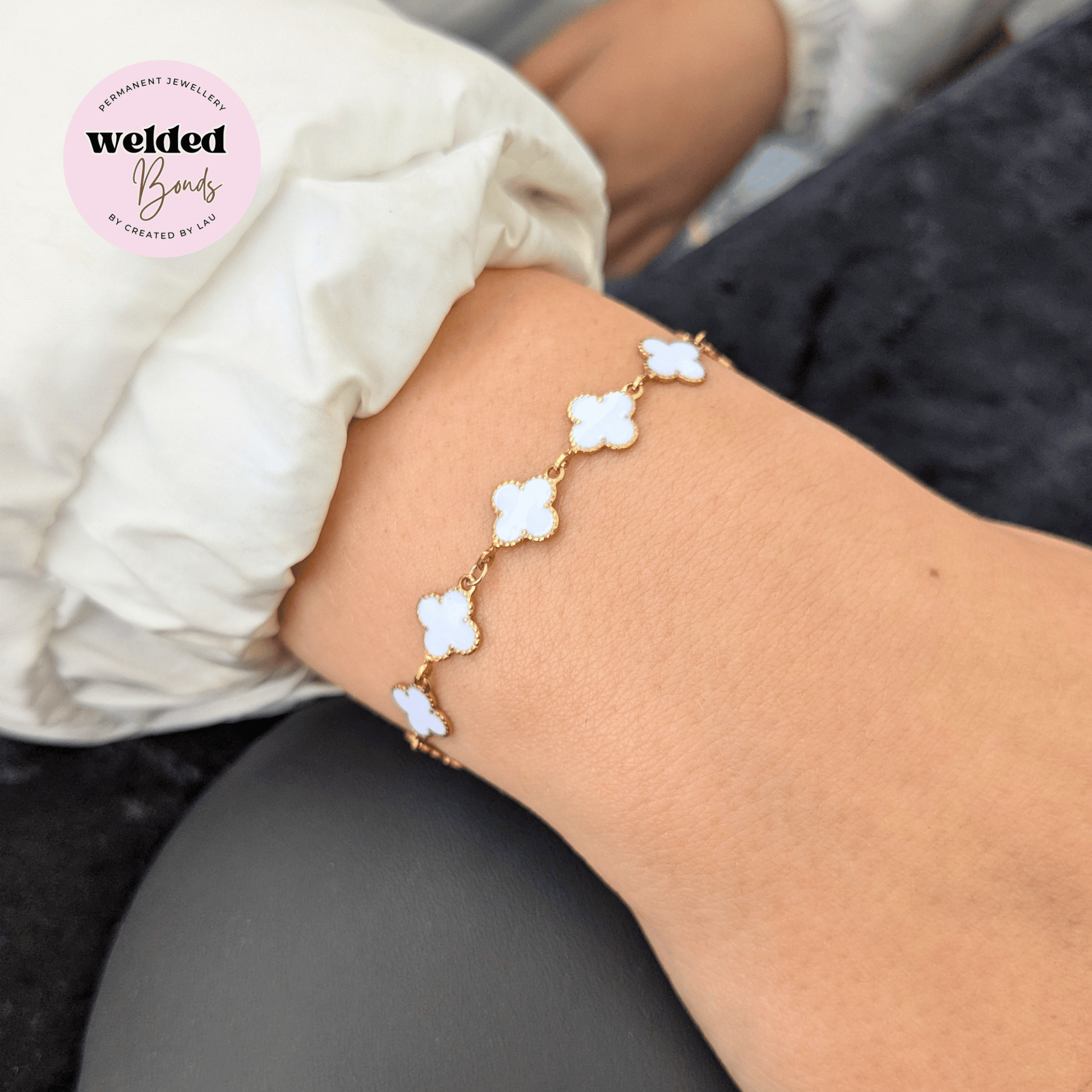 A gold-plated welded chain bracelet with five white clover-shaped charms, shown worn on a person's wrist. A pink circular logo for 'Welded Bonds' is visible in the top left corner.