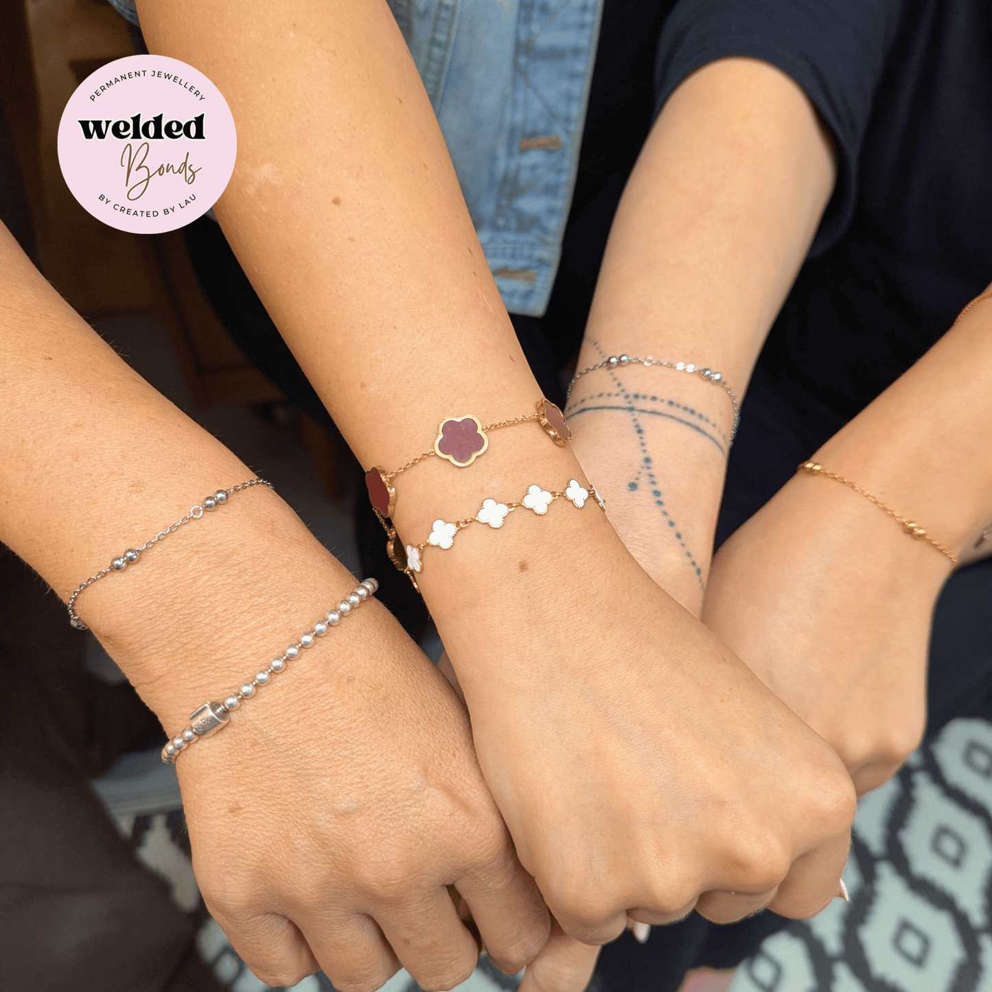 Three people's hands and wrists displaying different styles of welded bracelets. The bracelets include a silver beaded chain, a gold four-leaf clover with a burgundy center, and a gold chain with small white diamond charms.