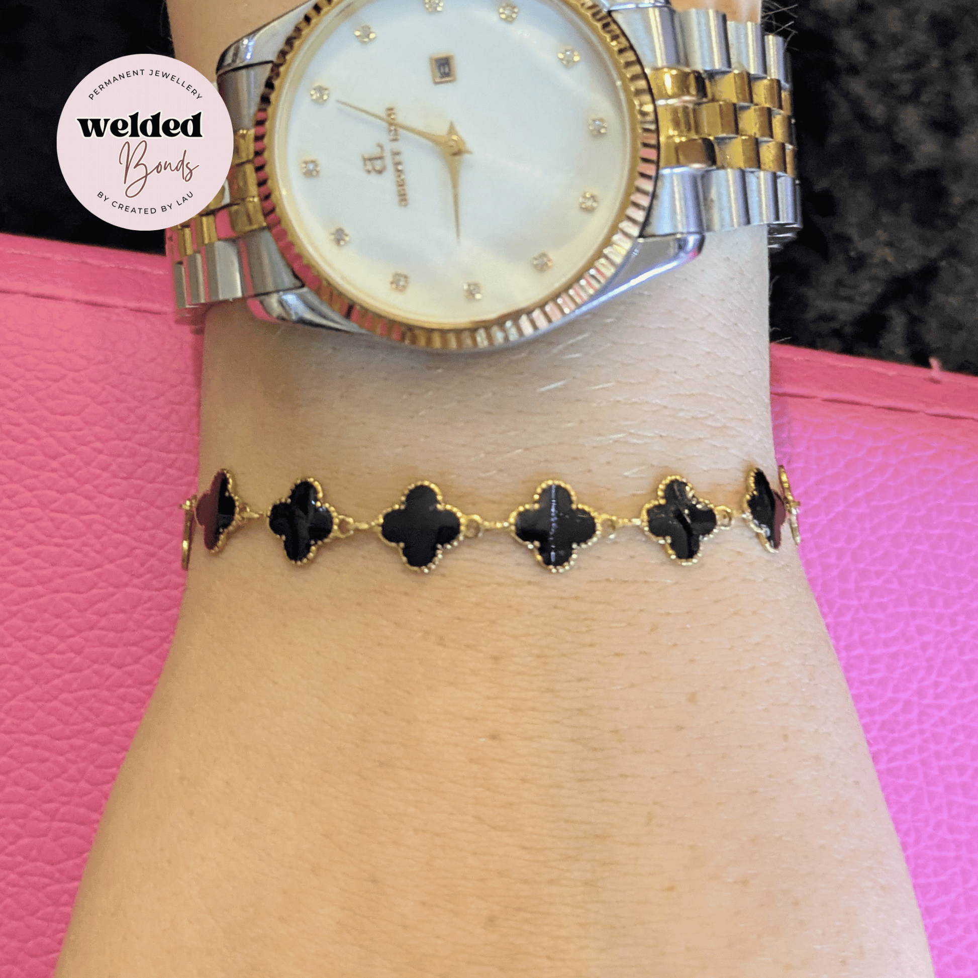A gold and silver welded bracelet with black clover motifs worn on a wrist below a gold and silver watch. A circular badge in the top left reads 'Welded Bonds'.