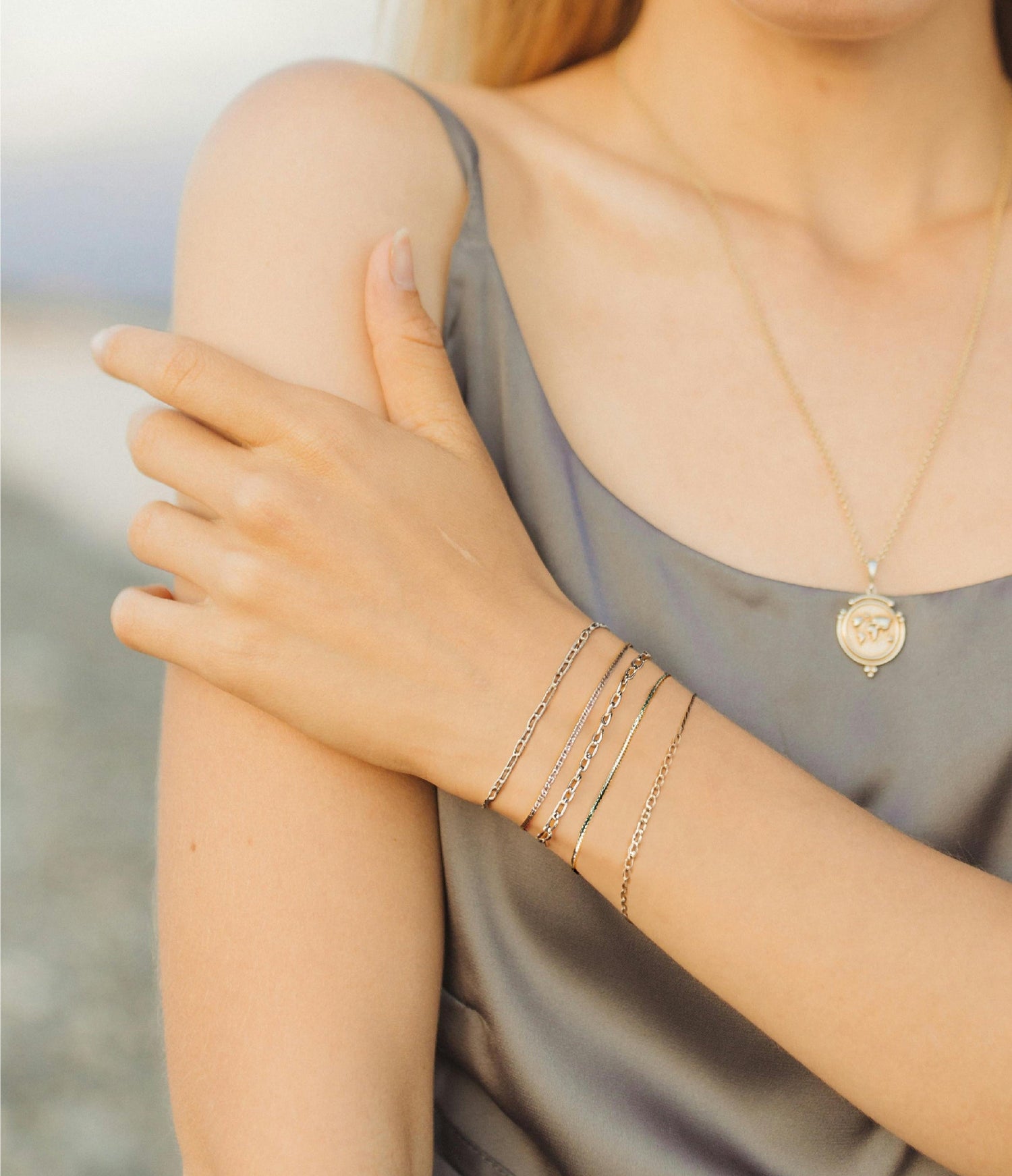 A woman wearing permanent gold jewellery, including a necklace, bracelet, and ring on a blurred beach background.