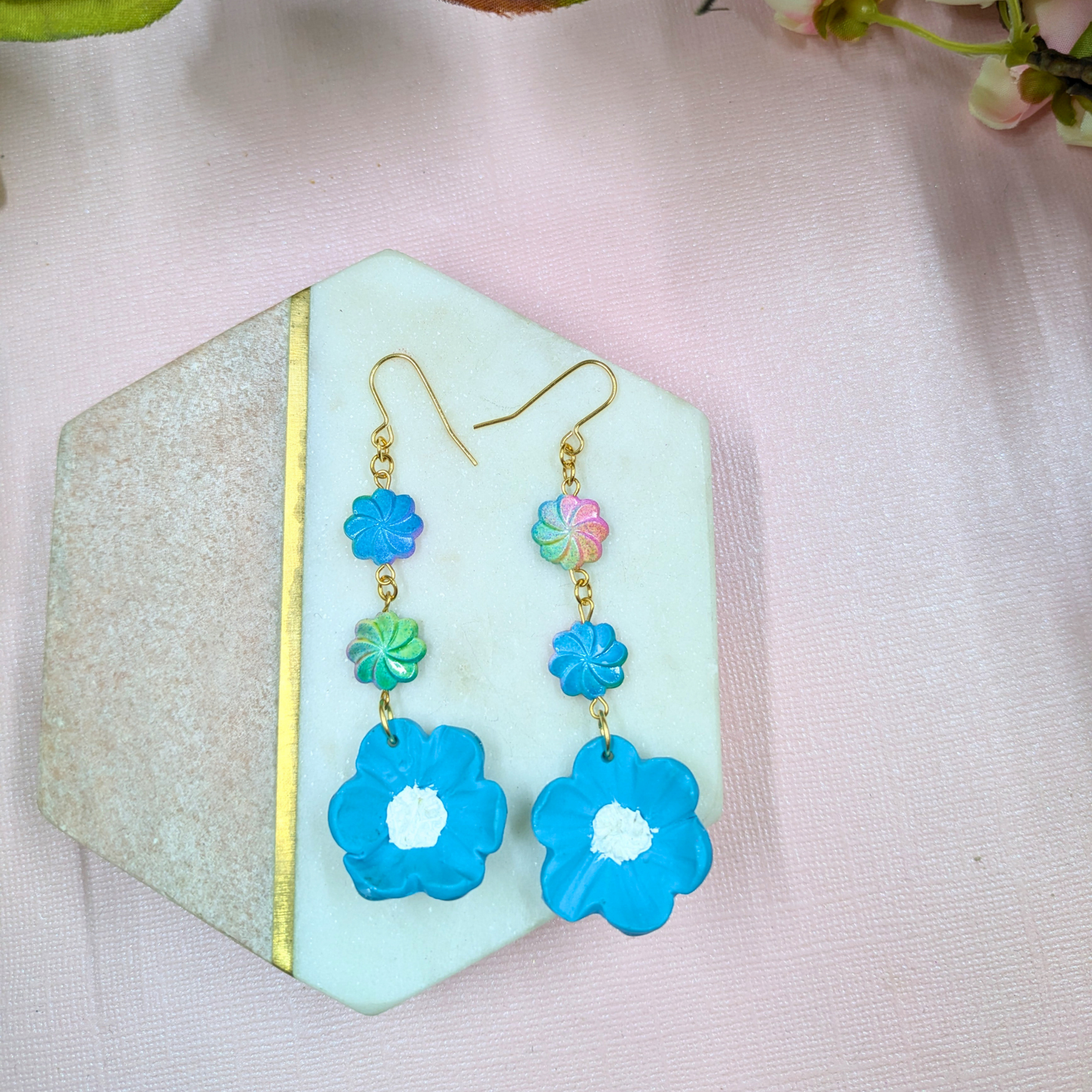 Blue flower-shaped Polymer Clay earrings wtih multi coloured recycled beads on a decorative stand with a soft pink background